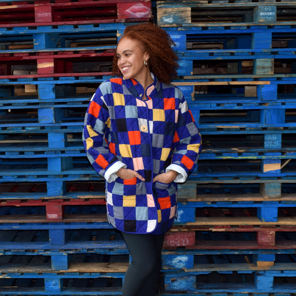 Woman in a colorful checkered quilted jacket standing between stacks of pallets.