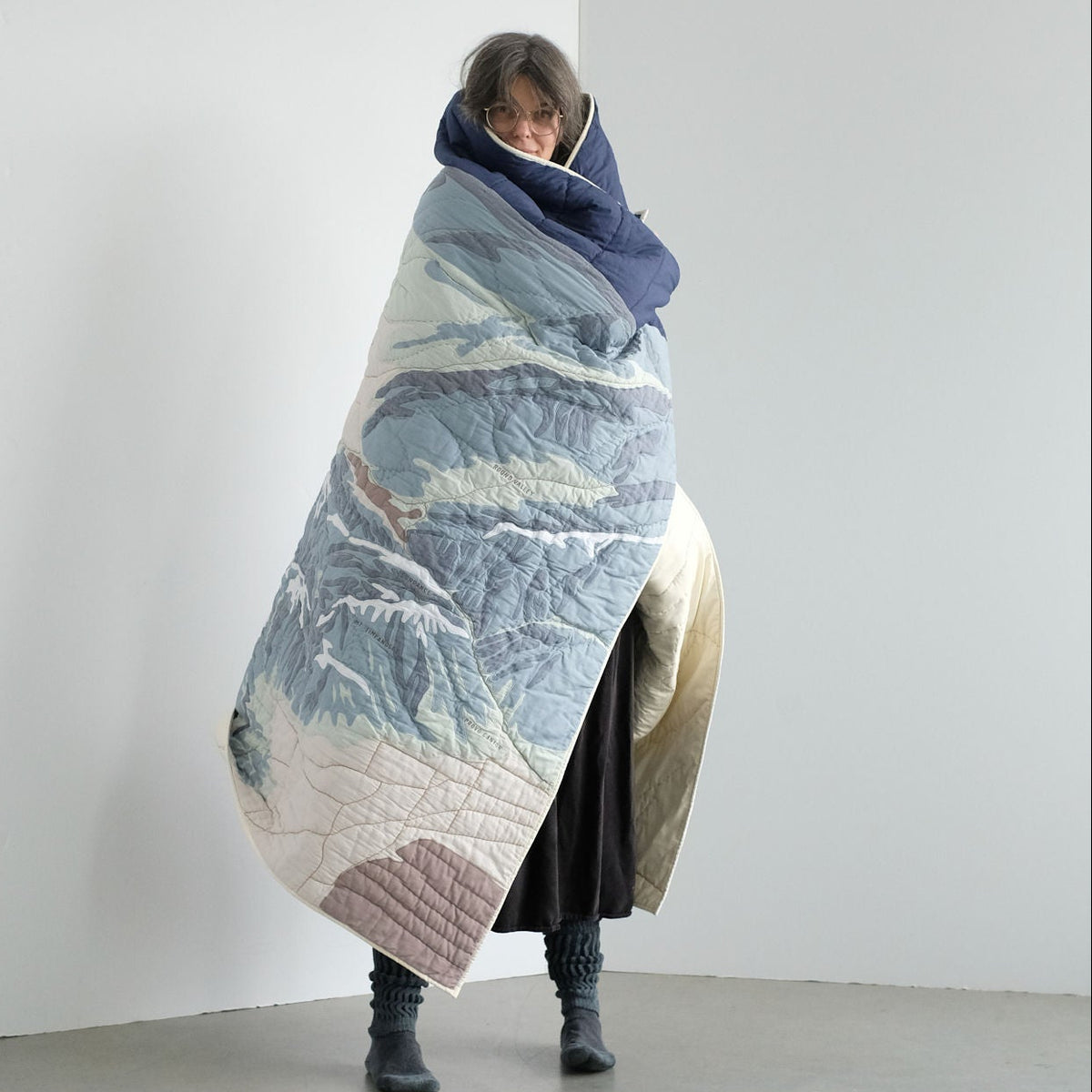 Person wrapped in a large, textured Wasatch Range quilt  against a plain white wall.