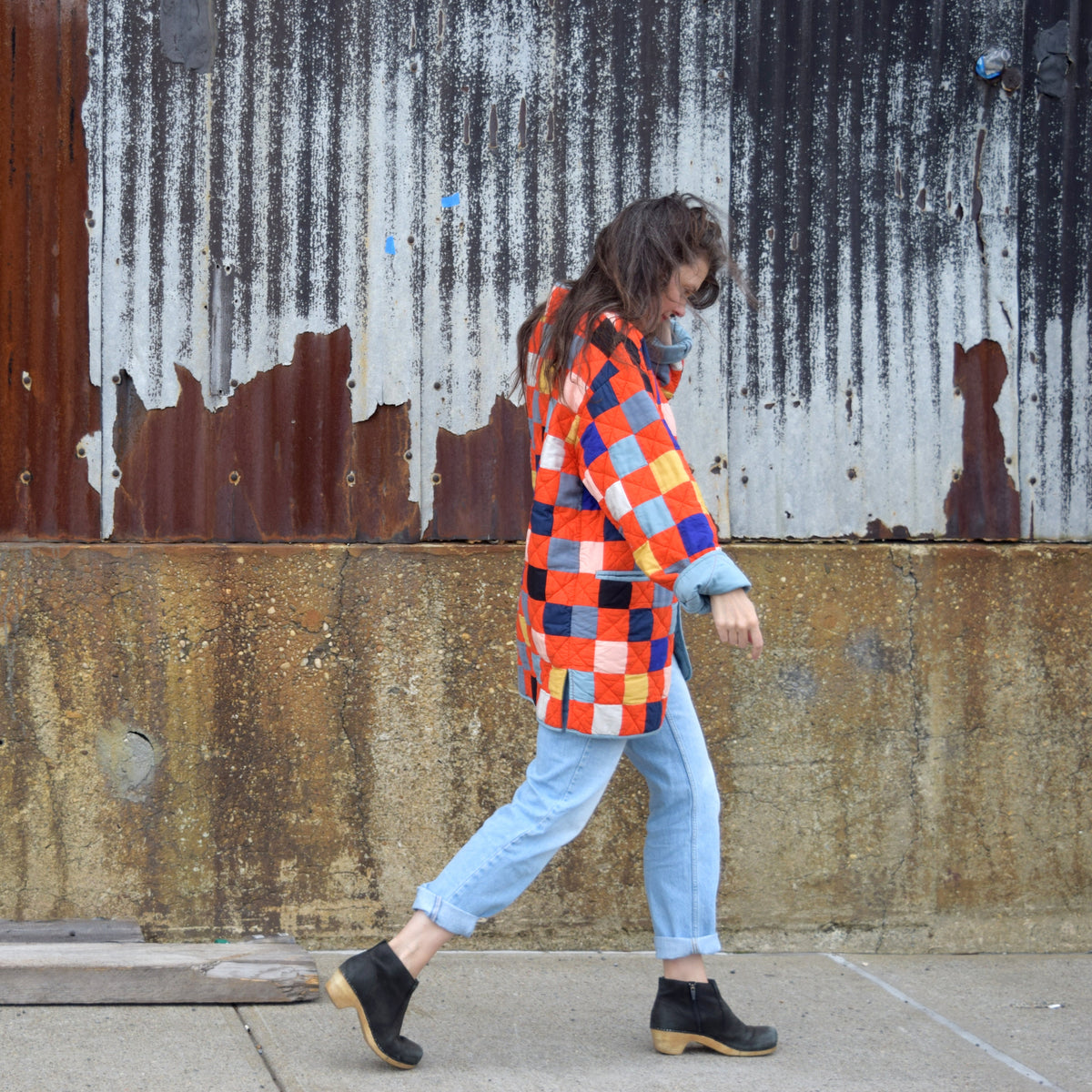 Person wearing a colorful checkered quilted jacket walking past a weathered wall.