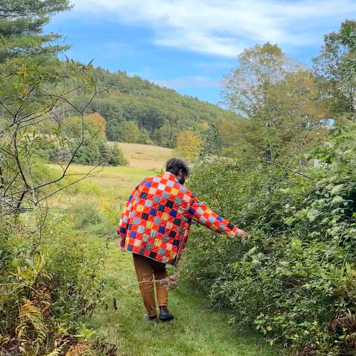 Person in a colorful quilted jacket walking along a trail in a forested area with trees and grass.